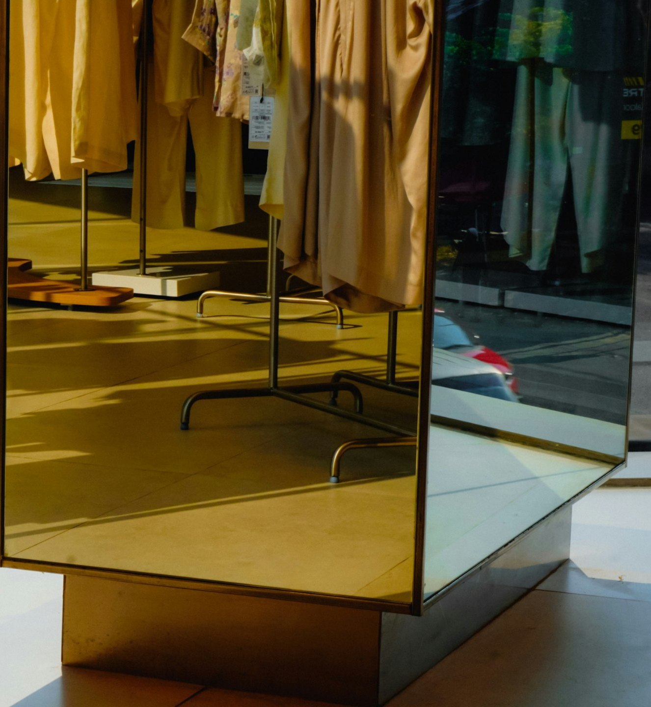 A mirrored display plinth in a boutique, reflecting rails of cream and sand garments in late afternoon light