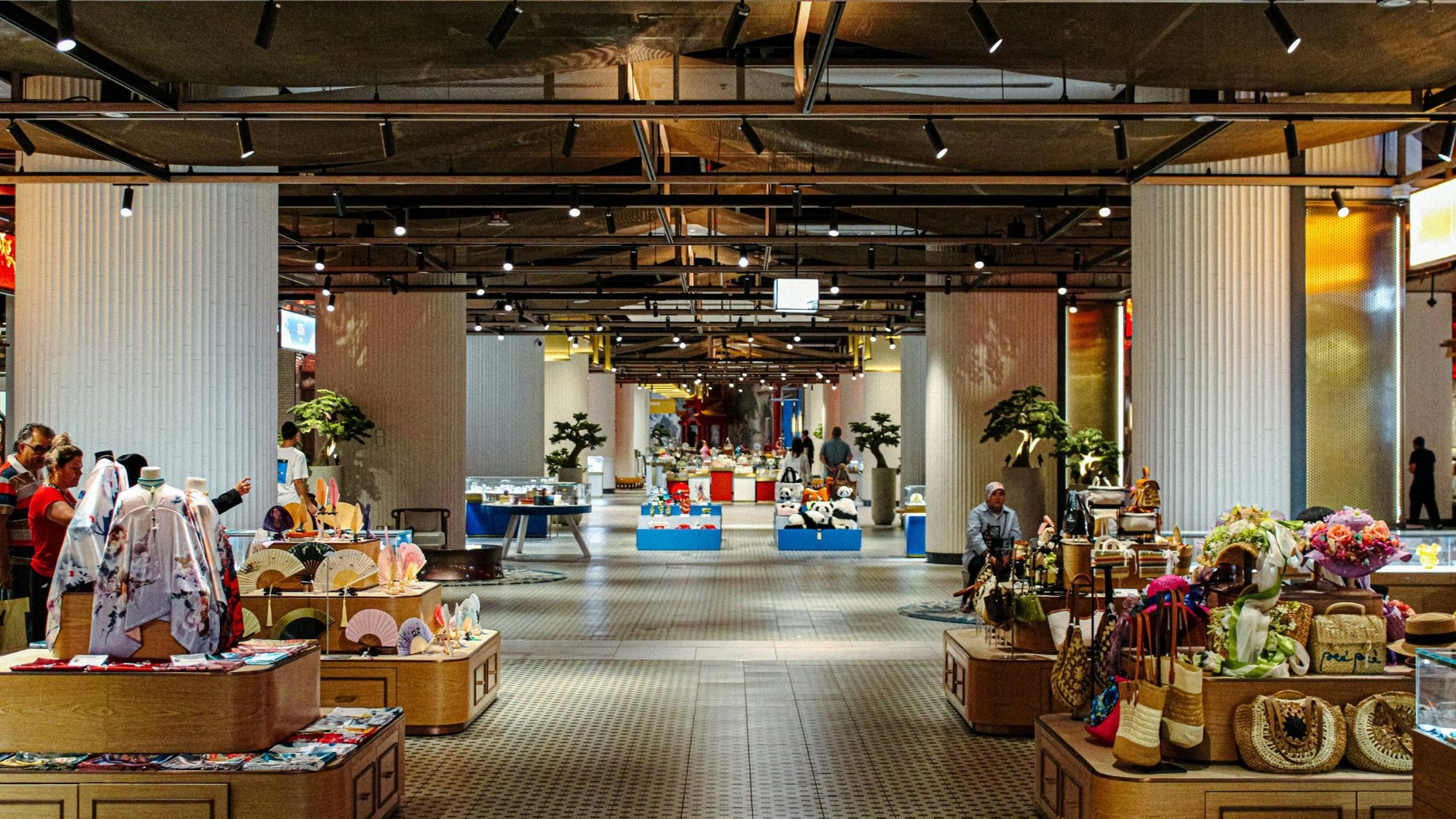 A long, light-filled retail hall with timber display tables of scarves, fans and woven bags