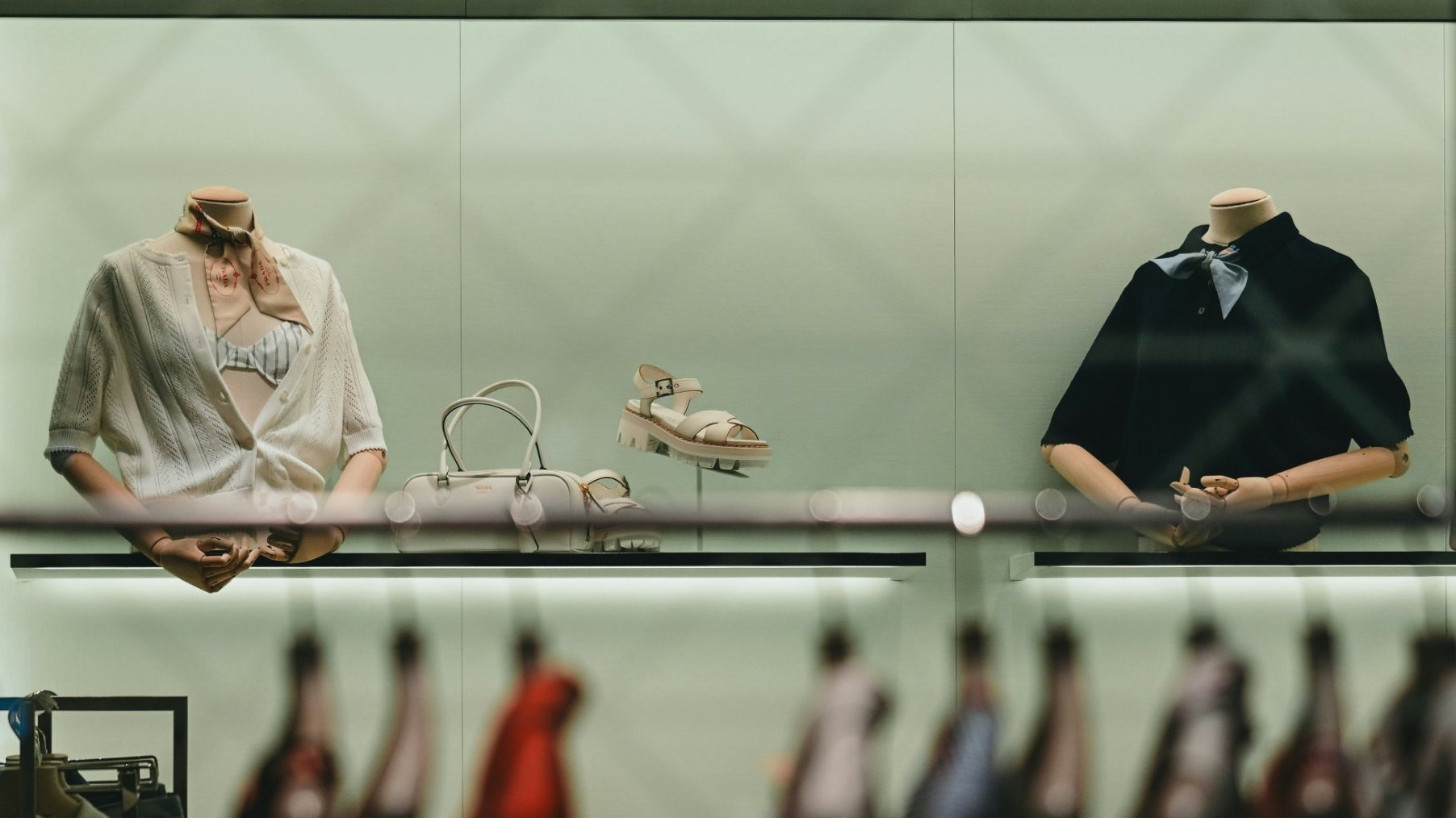 Two dressed forms on a lit shelf with a bag and sandals, rails of clothing soft in the foreground